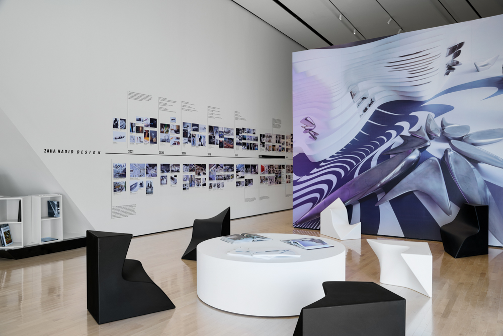 Image of exhibition with timeline of information, Zaha Hadid designed chairs, and large abstract photo mural